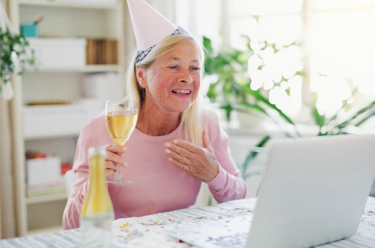 Senior woman with laptop, cake and wine indoors at home, celebrating distance birthday.