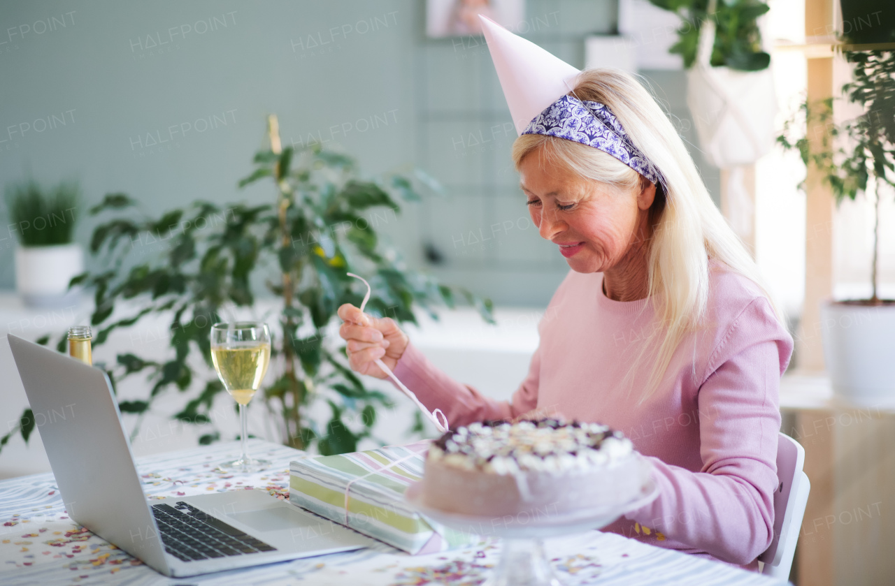 Senior woman with laptop, cake and wine indoors at home, celebrating distance birthday.