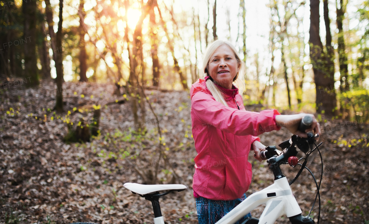 Active senior woman with bicycle standing outdoors in forest in nature. Copy space.