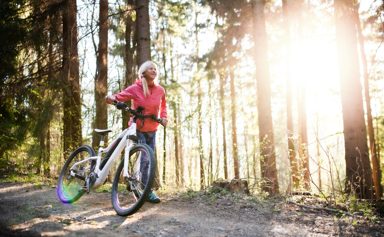 Active senior woman with e-bike cycling outdoors in forest in nature. Copy space.