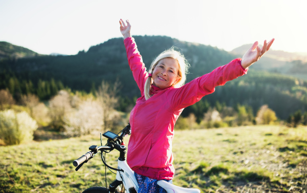 Front view of active senior woman with e-bike outdoors in nature, arms stretched.