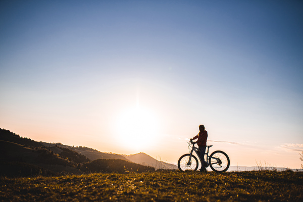 Side view of active senior woman with e-bike outdoors in nature at dusk. Copy space.