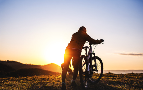 Rear view of active senior woman with bicycle outdoors in nature at sunset. Copy space.