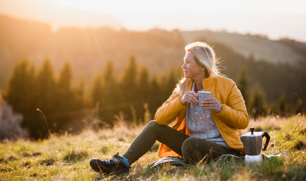 Attractive senior woman sitting outdoors in nature at sunset, relaxing with coffee. Copy space.