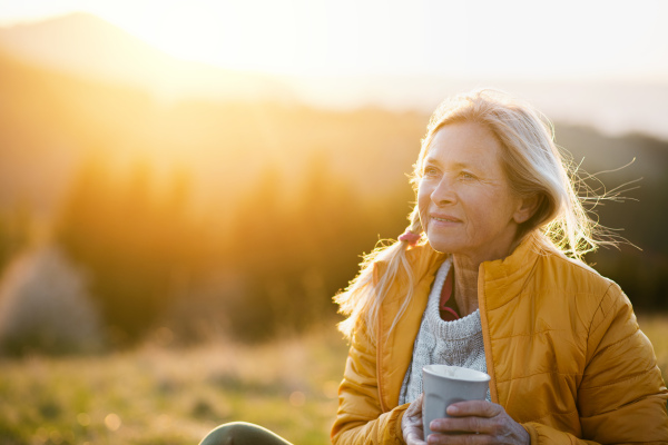 Attractive senior woman sitting outdoors in nature at sunset, relaxing with coffee. Copy space.