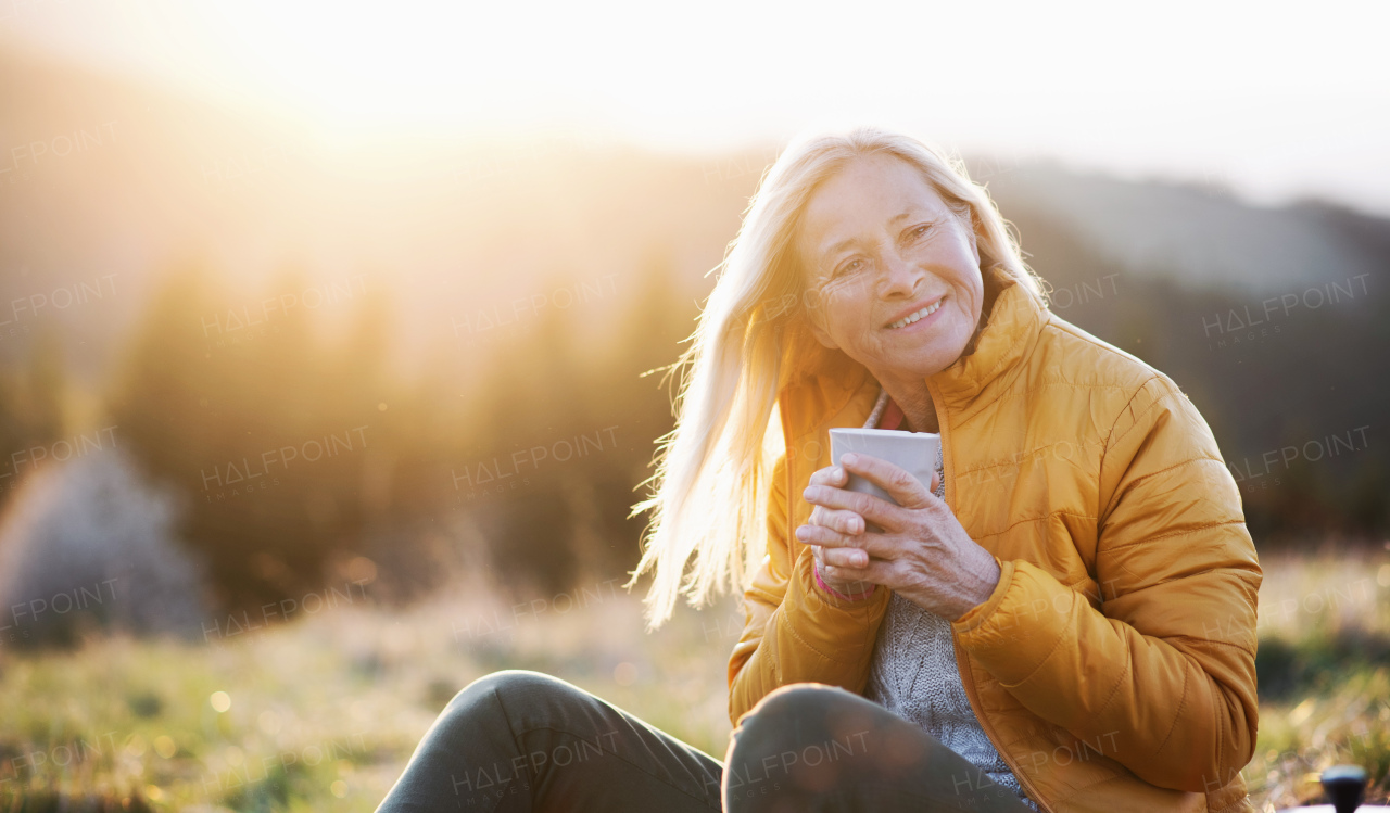 Attractive senior woman sitting outdoors in nature at sunset, relaxing with coffee. Copy space.