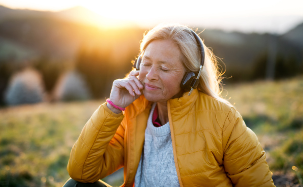 Attractive senior woman sitting outdoors in nature at sunset, relaxing with headphones.