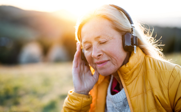 Attractive senior woman sitting outdoors in nature at sunset, relaxing with headphones.