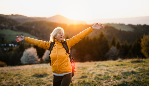 Front view of senior woman hiker standing outdoors in nature at sunset, arms stretched.