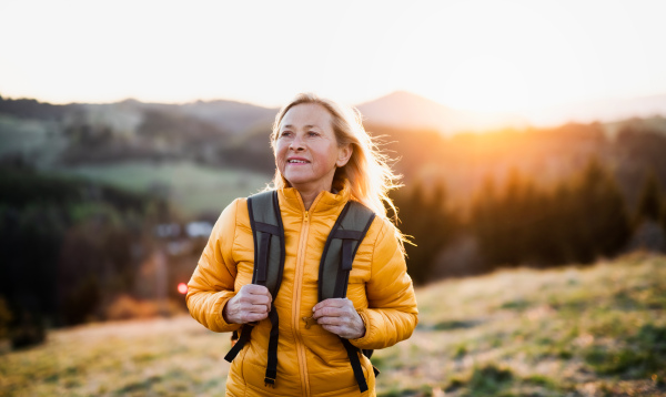 Attractive senior woman with backpack walking outdoors in nature at sunset, hiking.