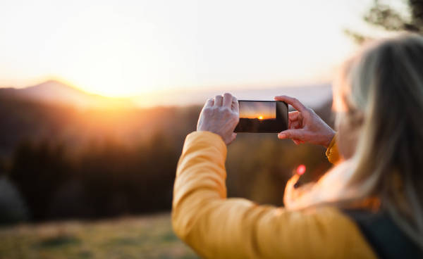 Senior woman hiker standing outdoors in nature at sunset, taking photograph with smartphone.