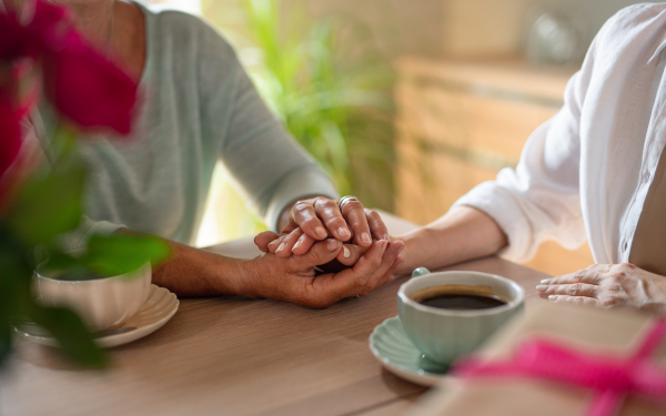 A close up of senior mother holding hand of adult daughter when having coffee together indoors at home