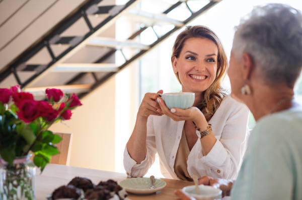 A happy mid adult daughter having coffee with senior mother indoors at home, sitting and talking.