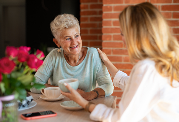 A happy senior mother talkinf with adult daughter when sitting and having coffee indoors at home.