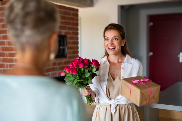 A happy adult daughter bringing gift and bouquet to senior mother indoors at home.