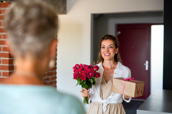 A happy adult daughter bringing gift and bouquet to senior mother indoors at home.