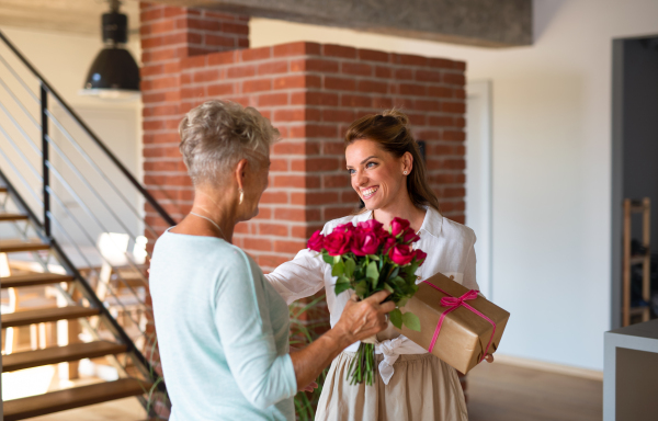 A happy adult daughter bringing gift and bouquet to senior mother indoors at home.