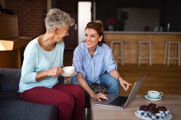 A happy senior mother having coffee with adult daughter indoors at home, sitting, talking and using tablet.