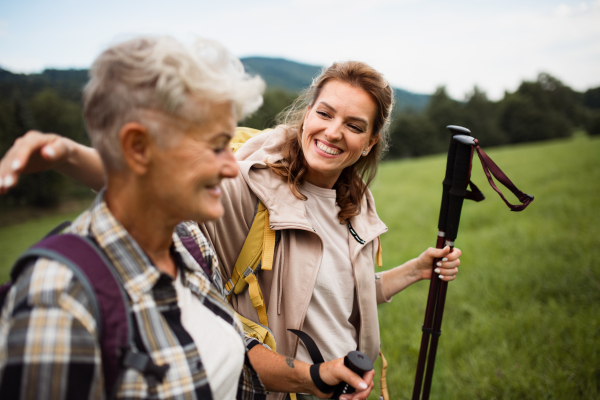 A happy mid adult woman with trekking poles hiking with active senior mother outdoors in nature.