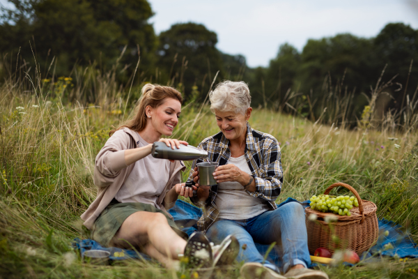 A happy senior mother and adult daughter sitting and having picnic outdoors in nature.