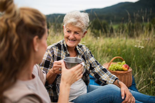 A happy senior mother and adult daughter sitting and having picnic outdoors in nature.