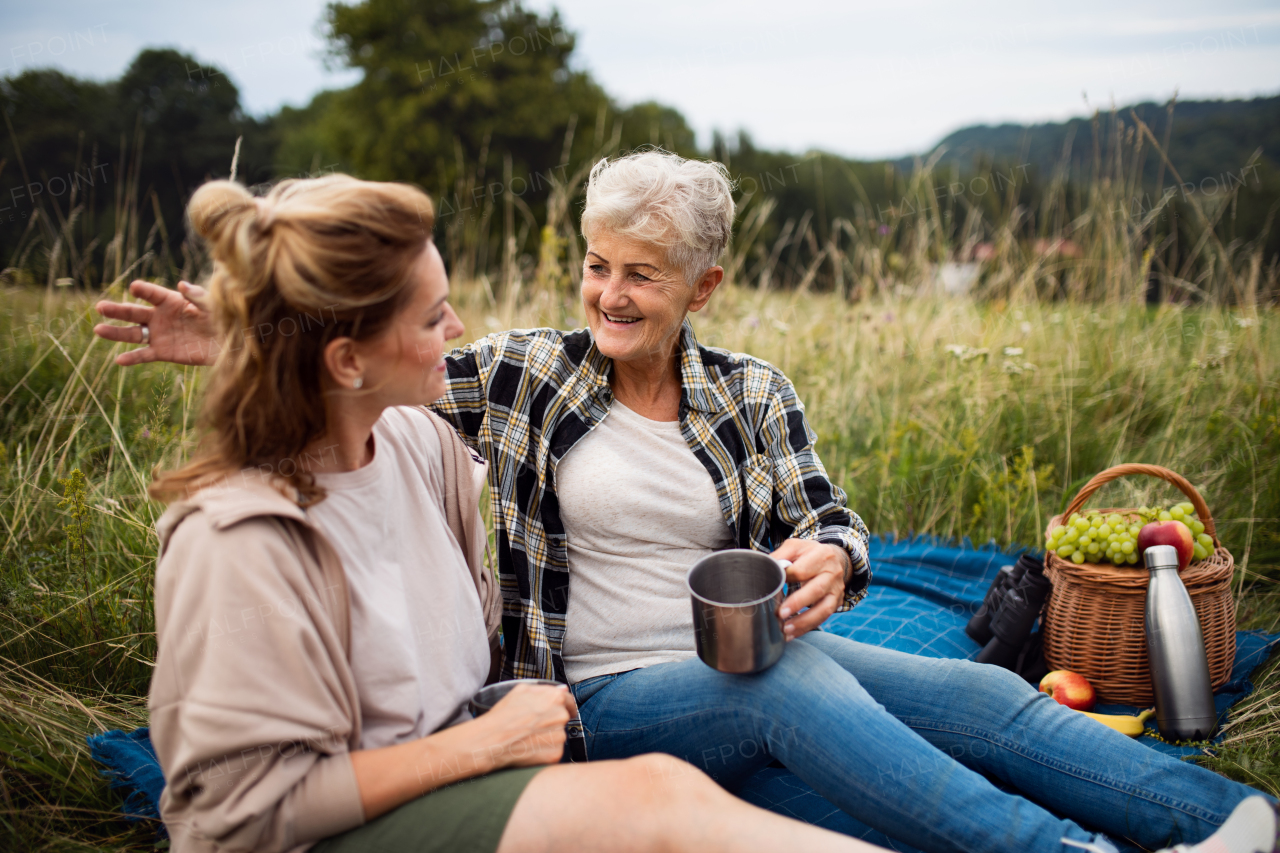 A happy senior mother and adult daughter sitting and having picnic outdoors in nature, talking.