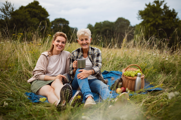 A happy senior mother and adult daughter sitting and having picnic outdoors in nature, looking at camera.