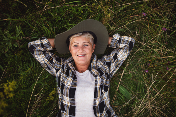 A top view of happy senior woman farmer lying on grass and looking at camera outdoors in meadow.