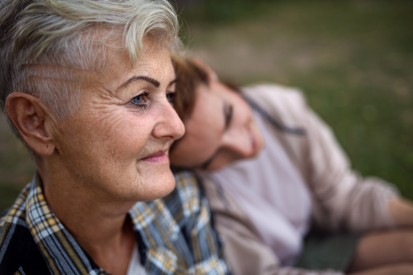 A close up of senior mother sitting on grass with adult daughter leaning her head on her outdoors.