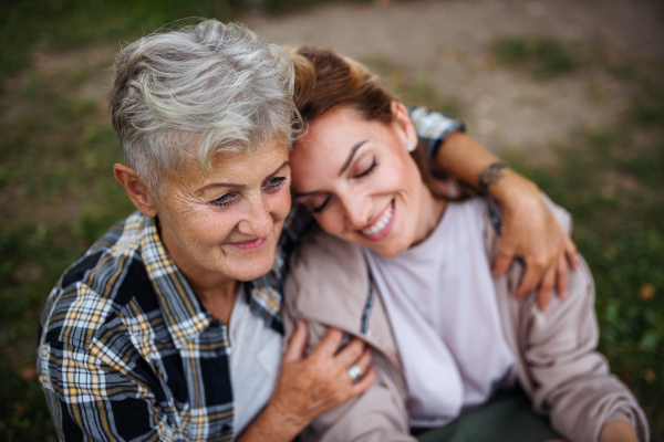 A close up of senior mother sitting on grass with adult daughter leaning her head on her outdoors.