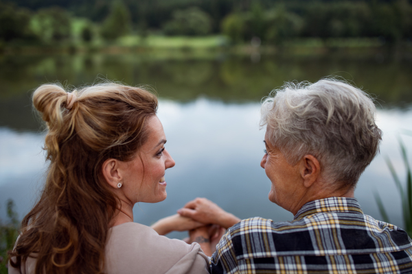 A rear view of happy senior mother embracing with adult daughter when sitting by lake outdoors in nature