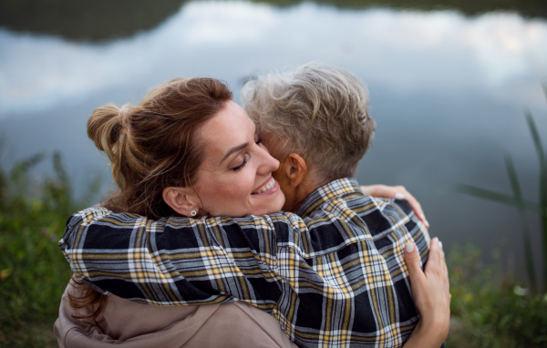 A rear view of happy senior mother embracing with adult daughter when sitting by lake outdoors in nature