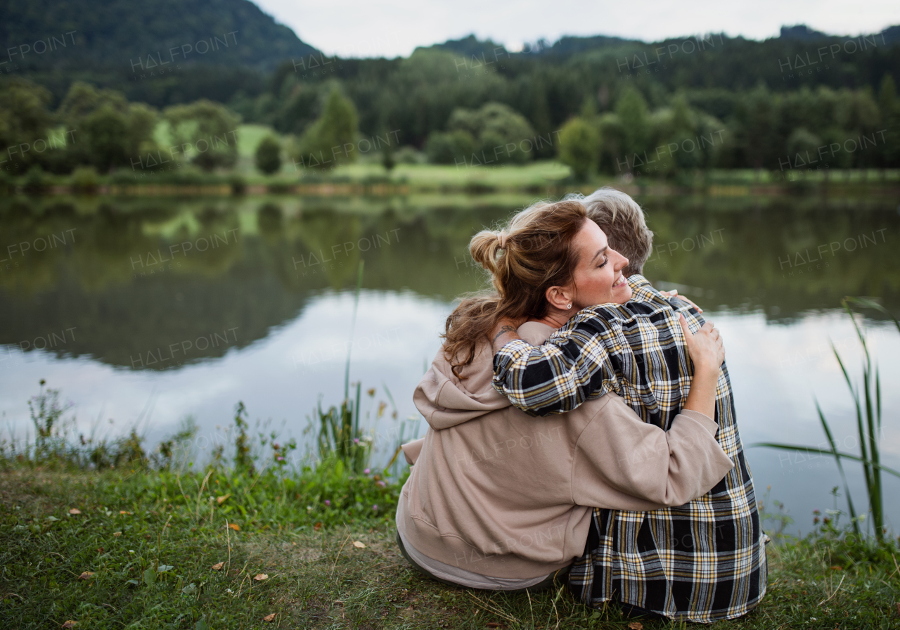 A rear view of happy senior mother embracing with adult daughter when sitting by lake outdoors in nature