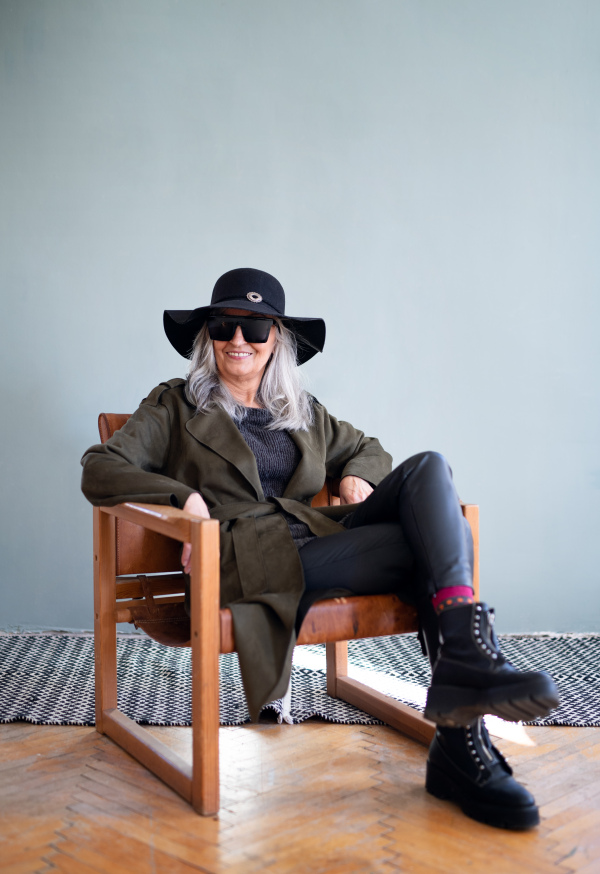 Front view portrait of senior woman with sunglasses and hat sitting indoors on chair, looking at camera.