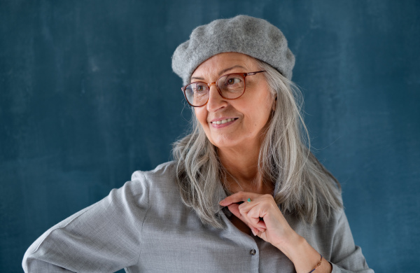 Studio portrait of senior woman with gray beret standing indoors against dark background.