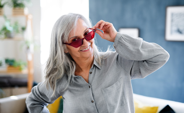 A portrait of senior woman with sunglasses sitting indoors on sofa at home, looking at camera.