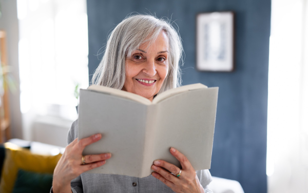 Portrait of senior woman with book indoors on sofa at home, looking at camera.