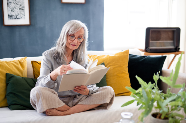 Portrait of senior woman sitting indoors on sofa at home, relaxing and reading book.