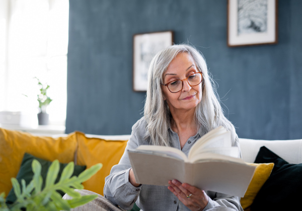 Portrait of senior woman sitting indoors on sofa at home, relaxing and reading book.