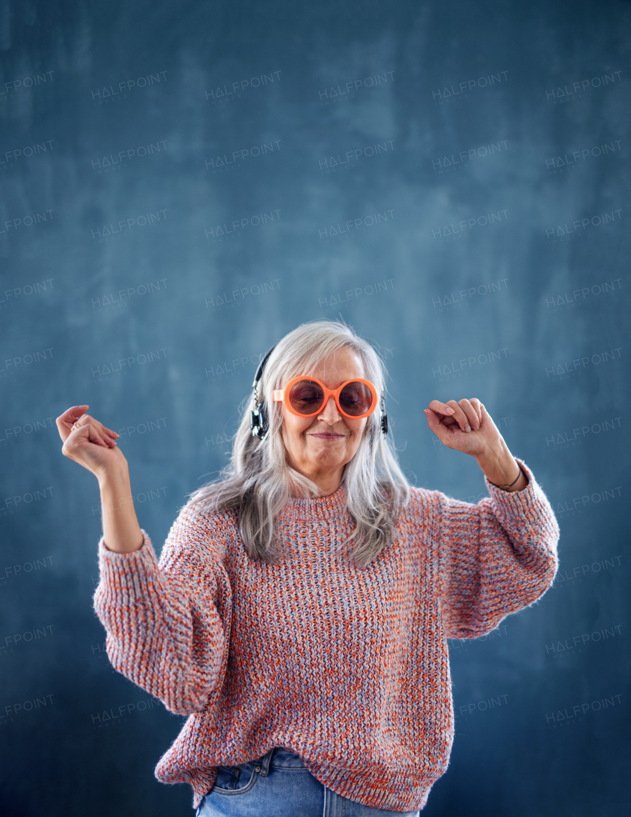 Portrait of senior woman with sunglasses and headphones standing indoors against dark background, dancing.