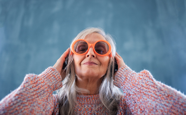 A portrait of senior woman with sunglasses standing indoors against dark background, listening to music.