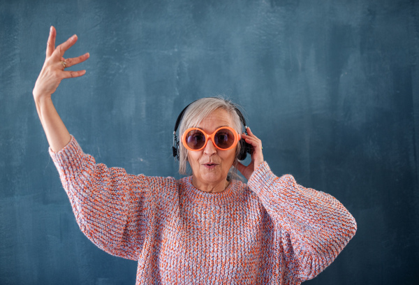 Portrait of senior woman with sunglasses and headphones standing indoors against dark background, having fun.