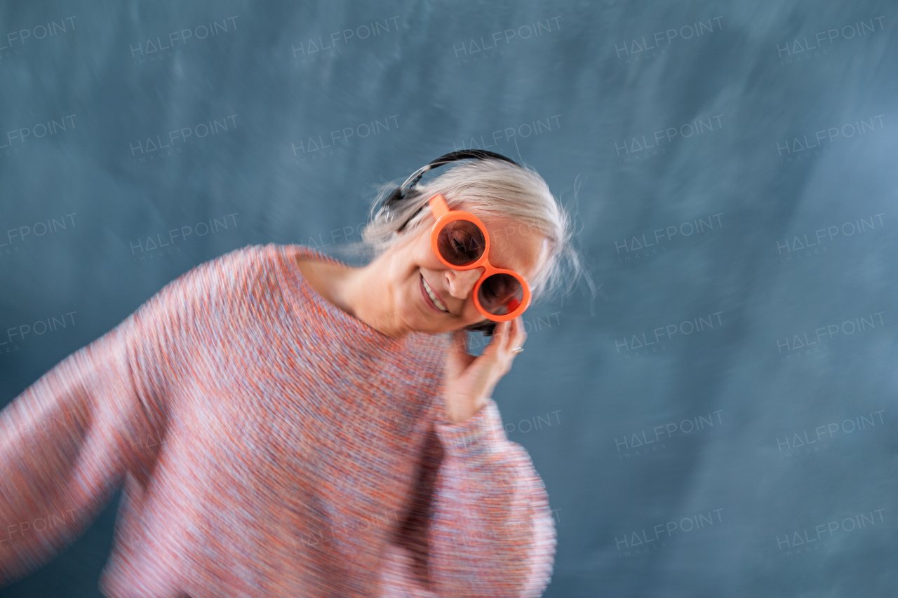 Portrait of senior woman with sunglasses and headphones standing indoors against dark background, motion blur.