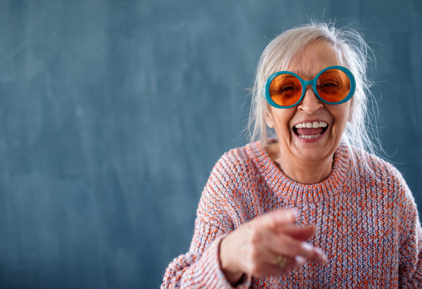 Portrait of senior woman with eccentric sunglasses standing indoors against dark background, laughing.