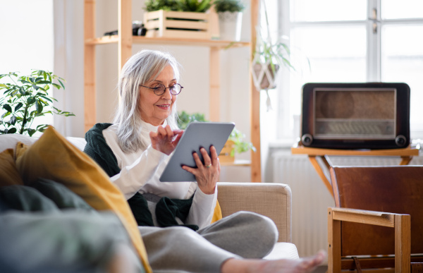 Portrait of senior woman sitting indoors on sofa at home, relaxing and using tablet.