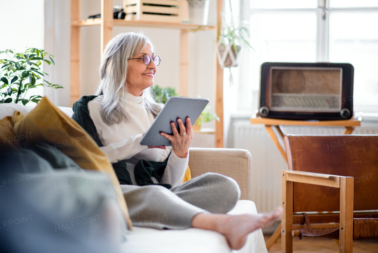 Portrait of senior woman sitting indoors on sofa at home, relaxing and using tablet.