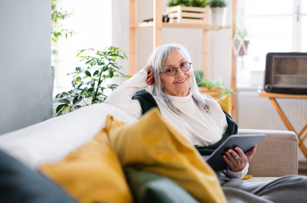 Portrait of senior woman sitting indoors on sofa at home, relaxing and using tablet.