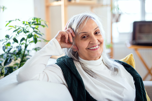 A portrait of senior woman sitting indoors on sofa at home, looking at camera.