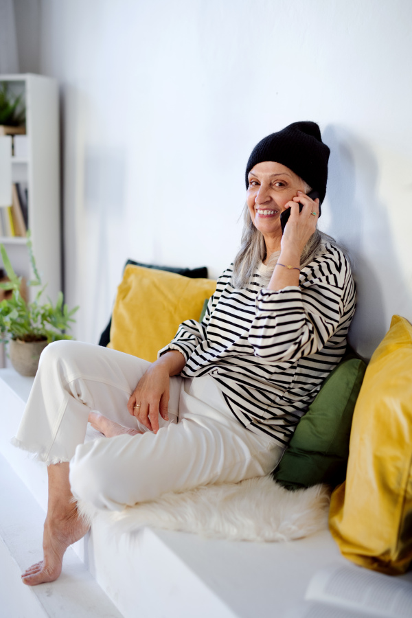 Portrait of senior woman with hat sitting indoors at home, using smartphone.