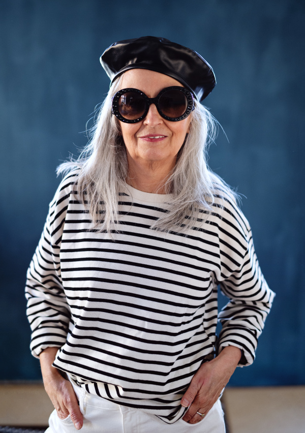 Studio portrait of senior woman with sunglasses and beret standing indoors against dark background.